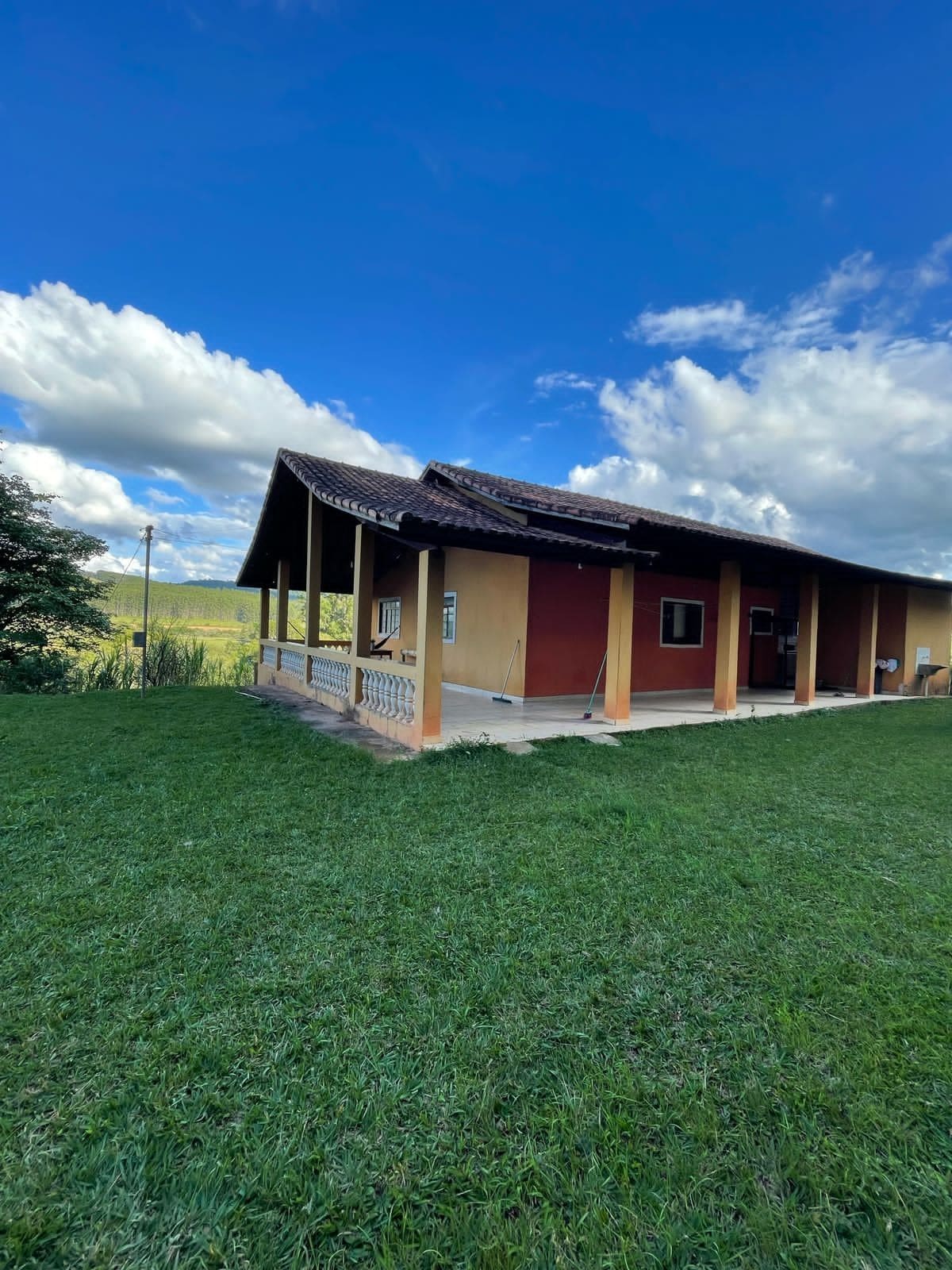 The exterior of the house is shown, featuring a broad porch with wooden pillars and a sturdy roof. The grassy area surrounding the home provides an inviting setting, while expansive blue skies with fluffy clouds add to the serene atmosphere.