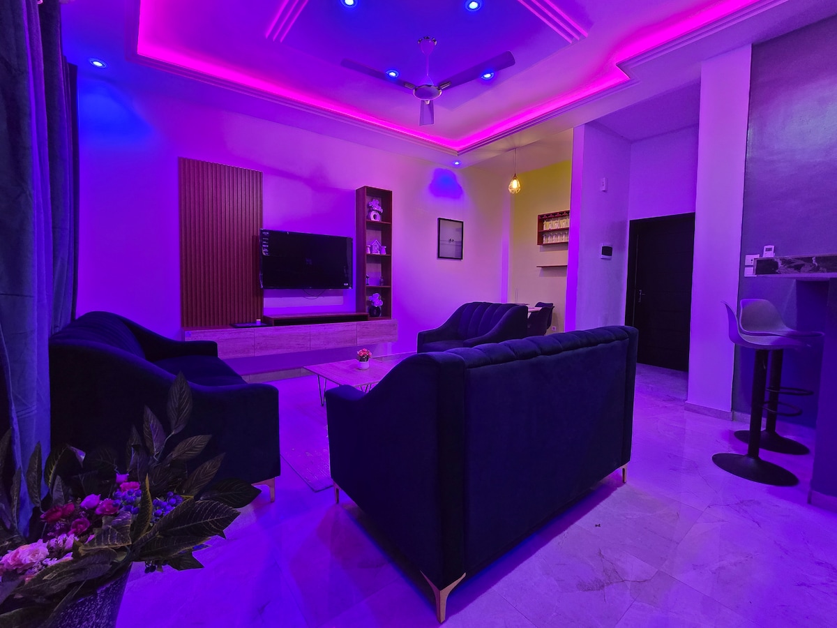 The living area features modern furnishings with plush seating arranged for comfort. A large television is mounted on the wall opposite. Soft lighting in purple hues enhances the ambiance, while a bar area is visible in the background, complemented by elegant decor.