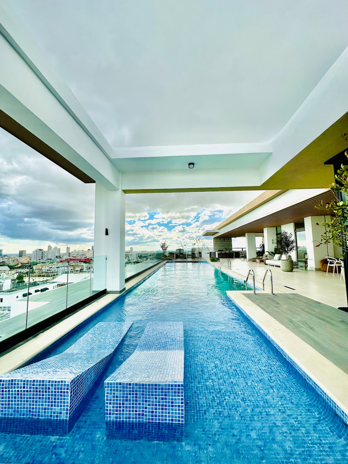 An outdoor infinity pool is presented, featuring comfortable lounge areas submerged at one end. The pool is surrounded by sleek tiles and glass railings, offering expansive views of the city skyline and sky, enhancing the sense of openness and tranquility.