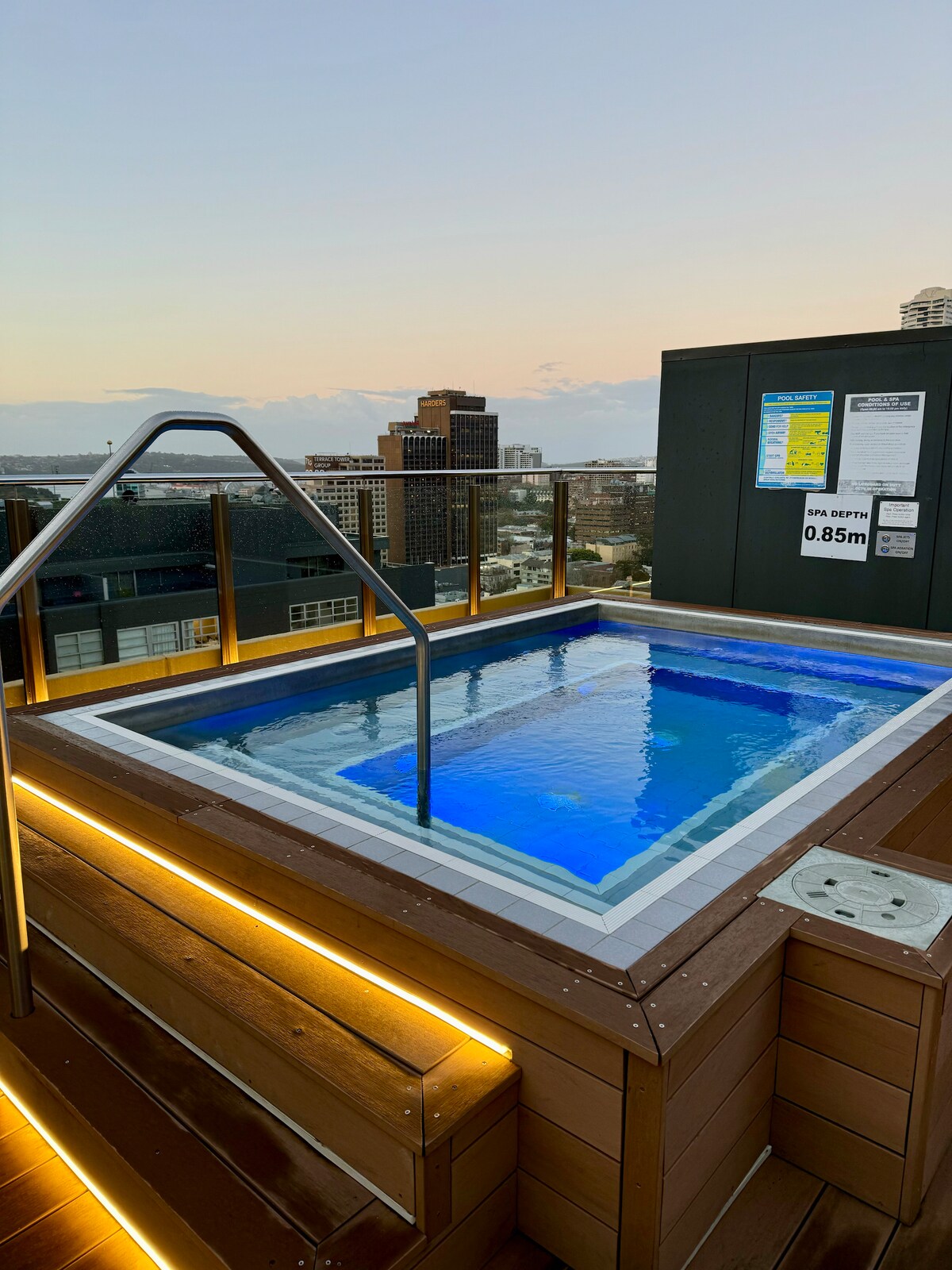 A rooftop spa area is showcased, featuring a rectangular spa pool with clear blue water. Surrounding the pool, wooden decking is illuminated by subtle lighting, enhancing the evening ambiance. A railing provides safety, and a city view is visible in the background.