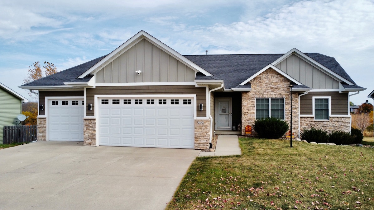 The exterior of a modern home presents a combination of light gray siding and stone accents. A spacious driveway leads to a two-car garage, showcasing multiple windows. The front yard features manicured grass and small shrubs, creating an inviting entryway.