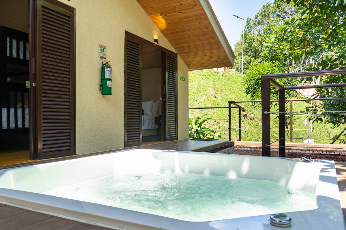 A private jacuzzi is set on a wooden deck, surrounded by lush greenery. Two rooms are visible beyond, featuring wooden shutters. The serene environment invites relaxation, with natural light filtering through the trees.
