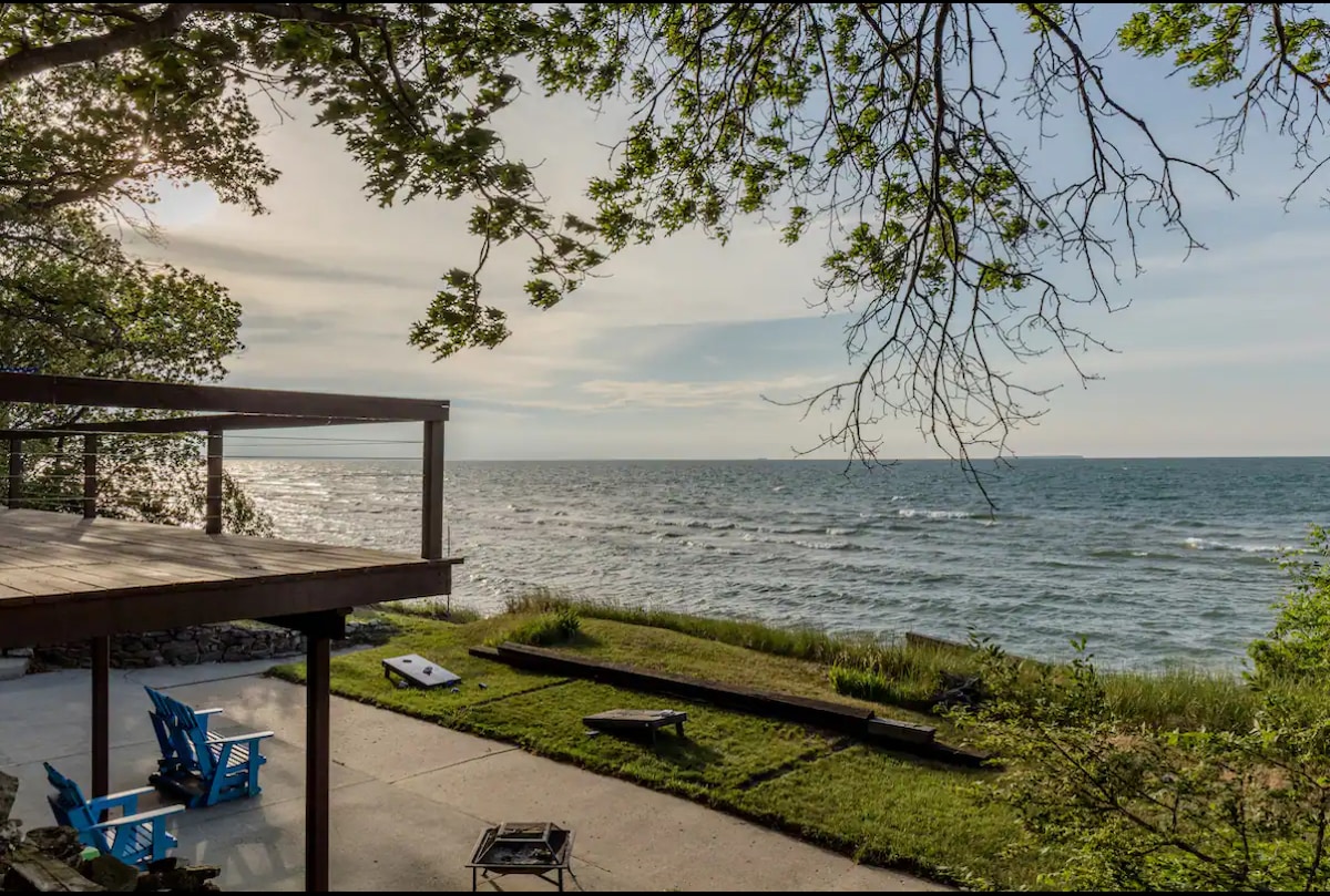 A peaceful view of Saginaw Bay is presented from a wooden deck. The calm water glistens under a bright sky, with waves gently lapping against the shore. Adjoining grass leads to the water's edge, framed by lush trees and simple outdoor seating.