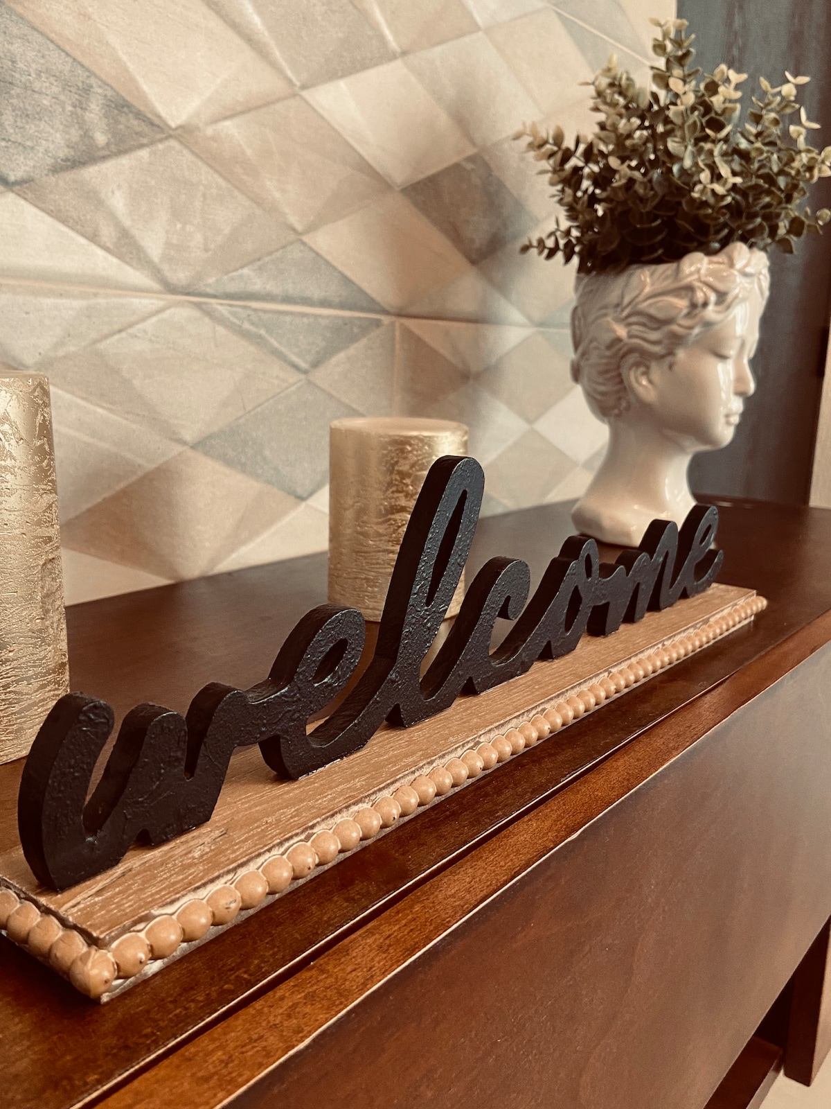 A decorative welcome sign in black metal is positioned on a wooden surface. Two cylindrical golden candle holders flank the sign, contributing to a warm atmosphere. A white bust sculpture and a small potted plant with greenery sit in the background against a textured wall.