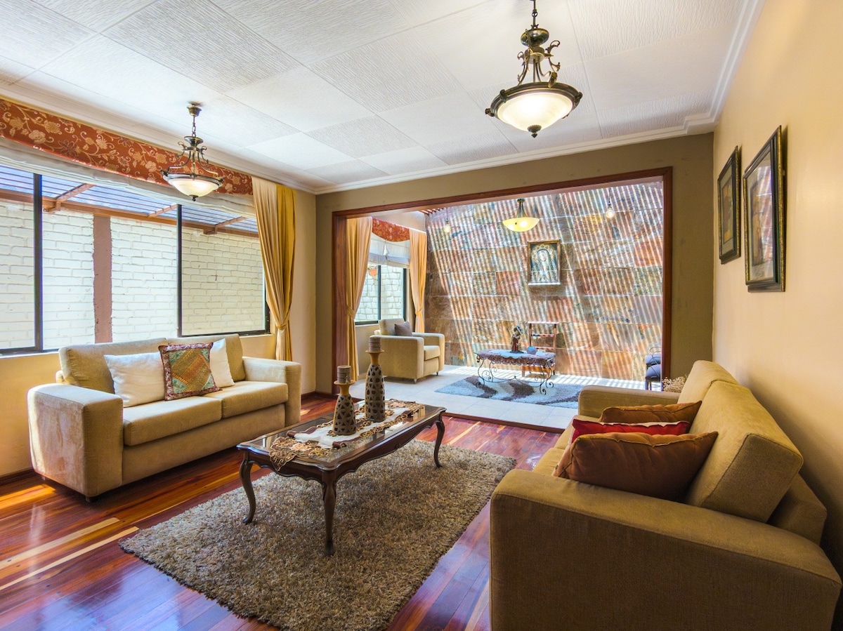 A spacious living area is furnished with two light-colored sofas and a coffee table. Large windows allow natural light to enter, framed by elegant curtains. A decorative rug adds warmth to the polished wooden floor. A view of the outdoor patio is visible through glass doors.