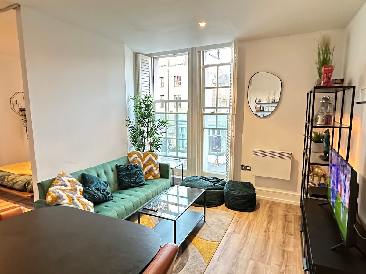 A bright living area features a comfortable green sofa, complemented by colorful cushions. A glass coffee table sits in front, surrounded by modern decor. Large windows allow natural light to fill the space, and a wall-mounted mirror adds depth. A Smart TV is positioned on a sleek entertainment unit.