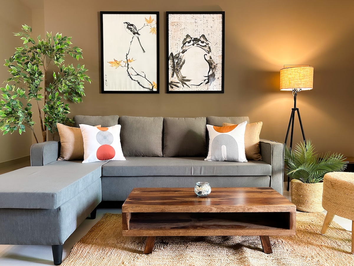 A spacious living area features a comfortable gray sofa accompanied by a stylish chaise lounge. Decorative pillows add subtle color, while two framed artworks are displayed on the wall. A wooden coffee table sits at the center, complemented by a woven rug and a soft-glow floor lamp.