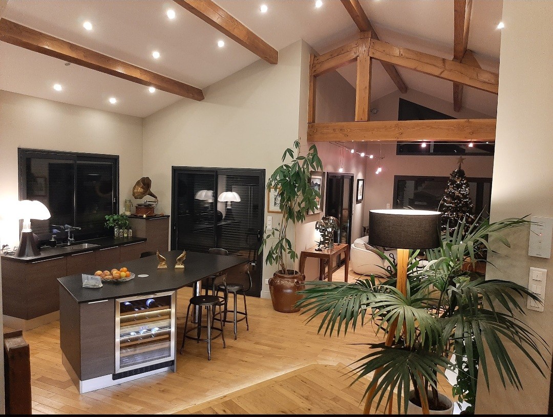 An open-concept kitchen features wooden beams and a large island with bar seating. A tropical plant adds greenery, while modern lighting enhances the spaciousness. Large windows filter in natural light, illuminating the warm wooden flooring and creating an inviting space.