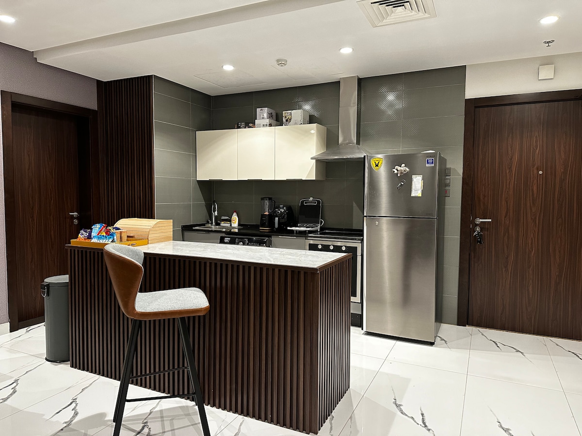 A modern kitchen features a sleek design with dark wood accents and light cabinetry. A central island with a stool provides seating. Appliances include a stainless-steel refrigerator and an oven. Cabinets are stocked with utensils, contributing to a fully equipped cooking space.