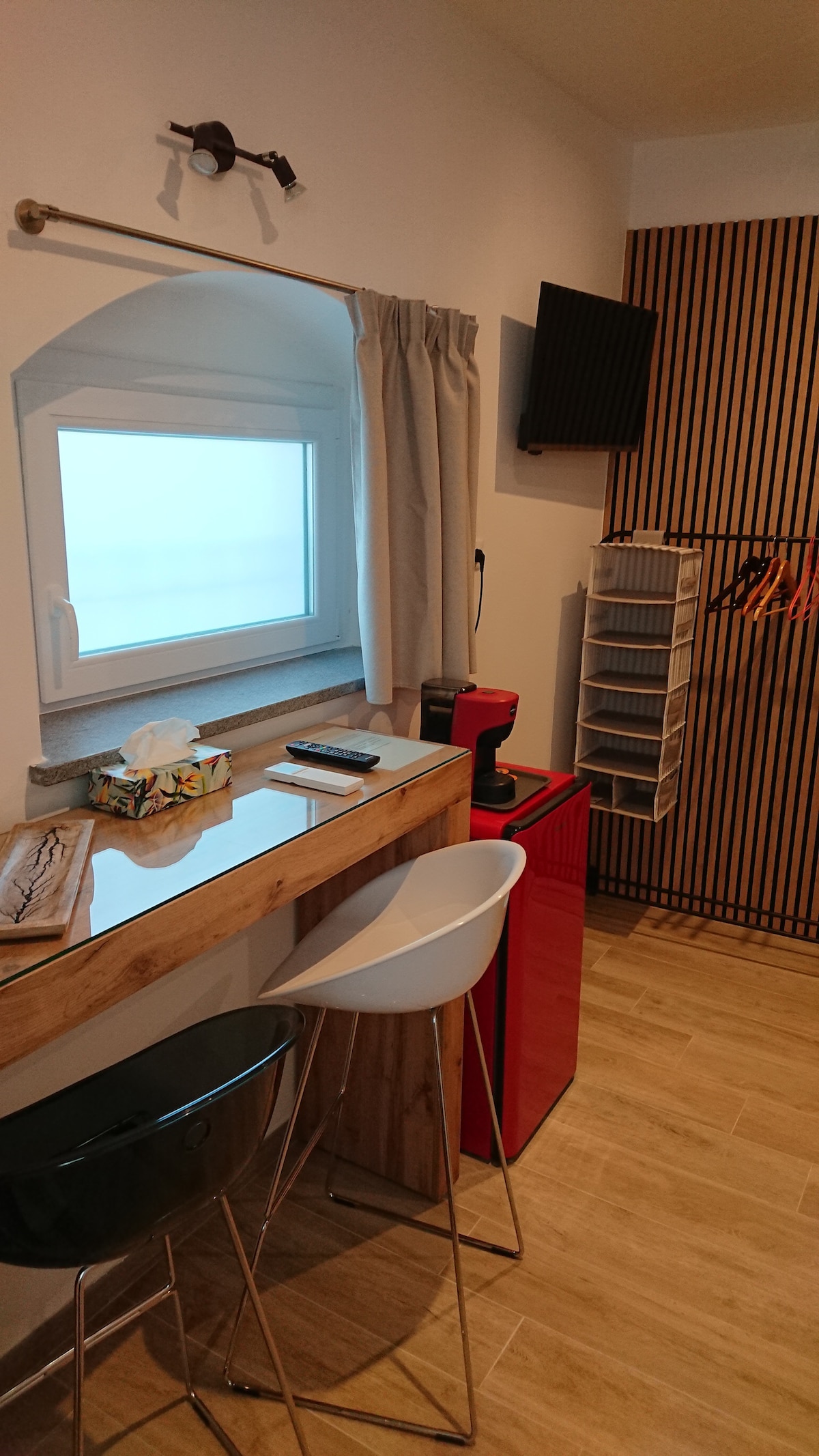 A small kitchenette area features a wooden counter with two stools, a coffee machine, and a red refrigerator. A wall-mounted TV is positioned above the counter. Natural light is filtered through a window covered by light curtains, and a hanging organizer is visible nearby.