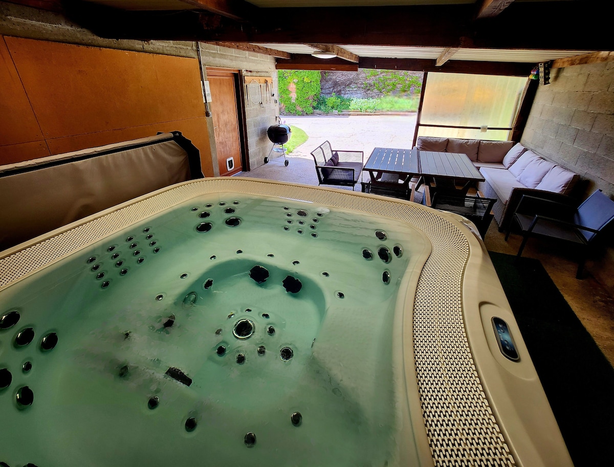 A seven-person spa is positioned under a covered area, surrounded by a seating arrangement with a couch. A table is also visible nearby, creating a relaxed atmosphere. Natural light filters in through large openings, enhancing accessibility to the outdoor area.