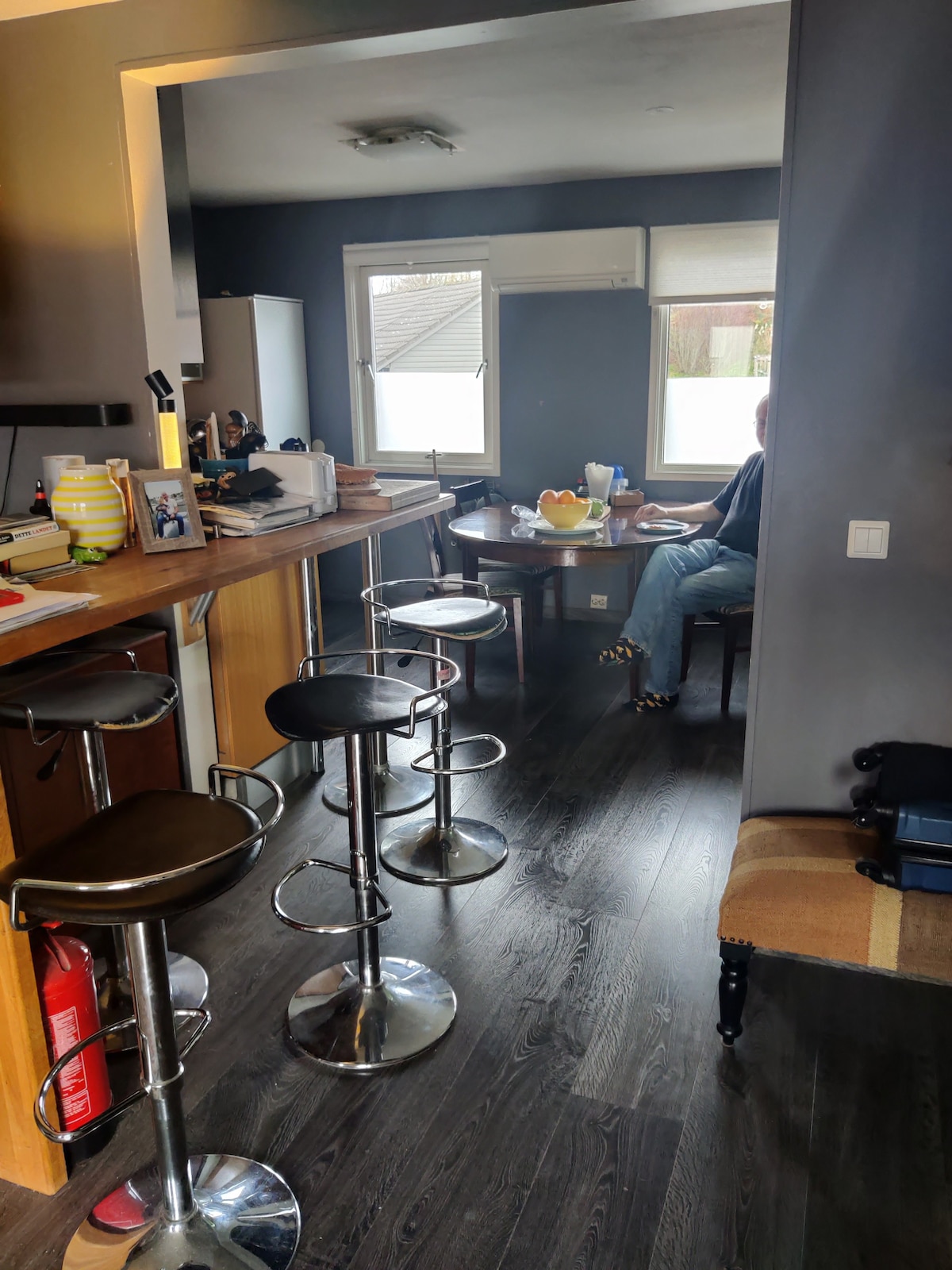 A modern kitchen area features sleek bar stools beside a wooden countertop. Natural light enters through a large window, illuminating the space. The room is equipped with a refrigerator and various kitchen items, providing a functional and inviting environment.