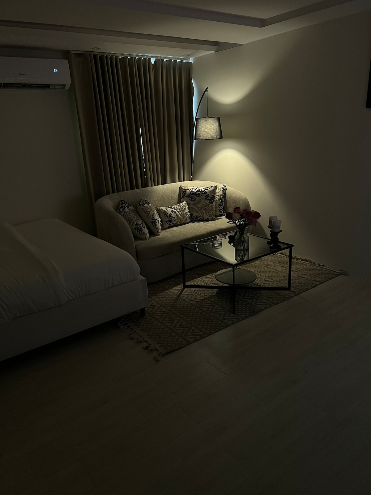 A cozy seating area is presented, featuring a light-colored sofa adorned with decorative pillows. A glass coffee table is centered on a patterned rug, complemented by a lamp providing soft illumination. The adjacent bed is elegantly dressed with white linens, enhancing the serene environment.