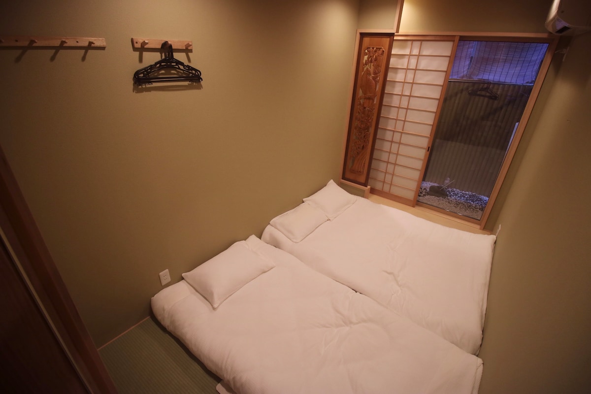 A minimalist bedroom features two spacious futons placed on tatami flooring. A wooden rack with hangers is positioned on the wall beside a shoji screen window, allowing soft natural light to filter in.