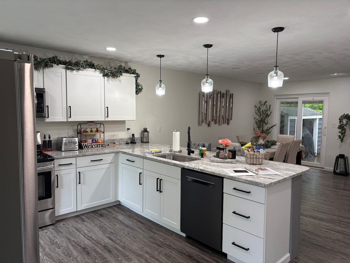 A modern kitchen is displayed, featuring white cabinetry and a granite countertop. Pendant lights illuminate the space, highlighting the open layout. A dining area is visible in the background, with a view of an outdoor space through a glass door.
