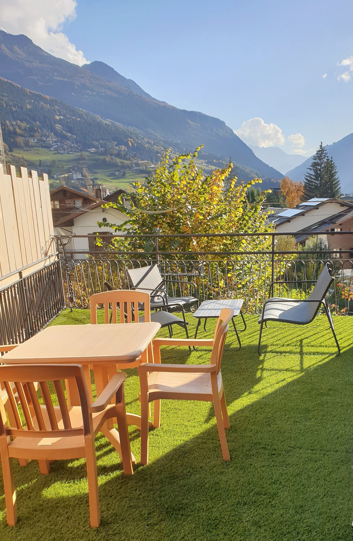 An outdoor terrace features a table with four wooden chairs, surrounded by two lounge chairs. Lush, green grass covers the flooring. The backdrop includes mountains and trees, reflecting natural colors under a bright sky.