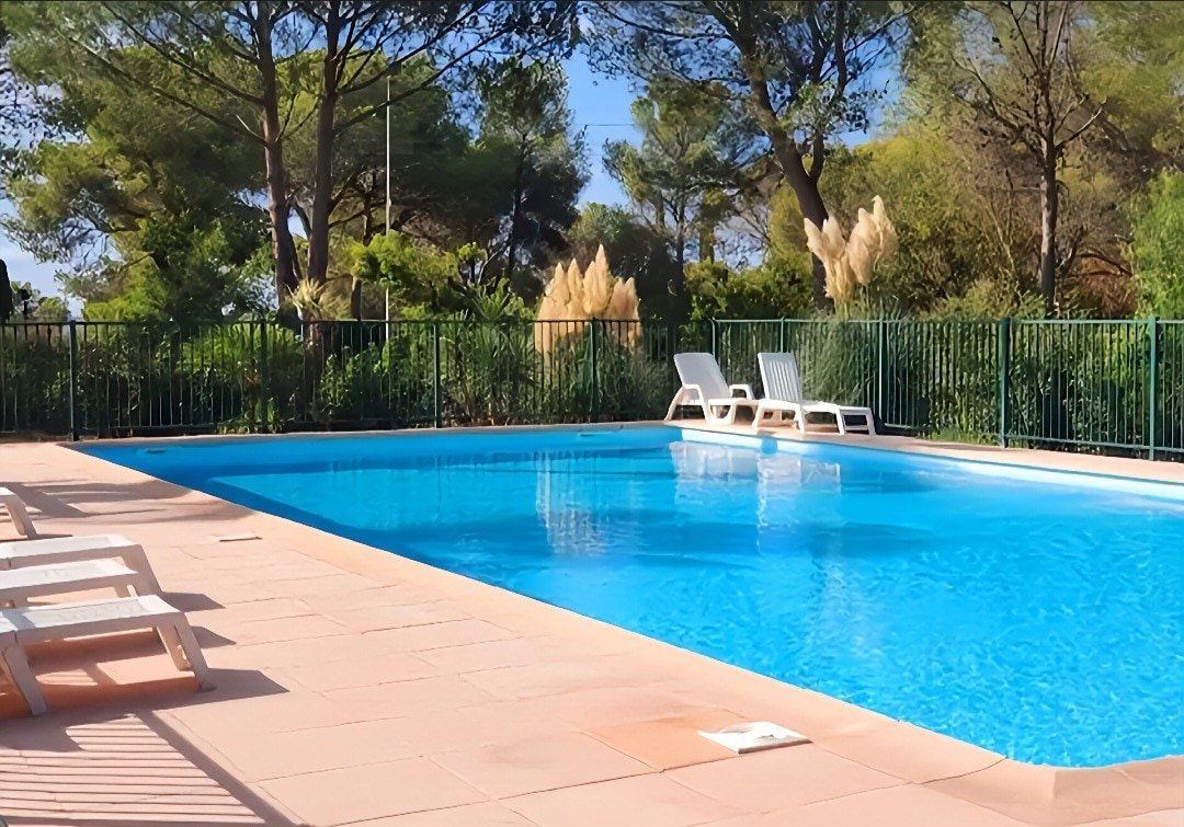 The image displays a clear, inviting swimming pool surrounded by well-maintained greenery and tall trees. Two white lounge chairs are positioned near the edge of the pool on a spacious tiled area, providing a peaceful relaxation spot in a tranquil setting.