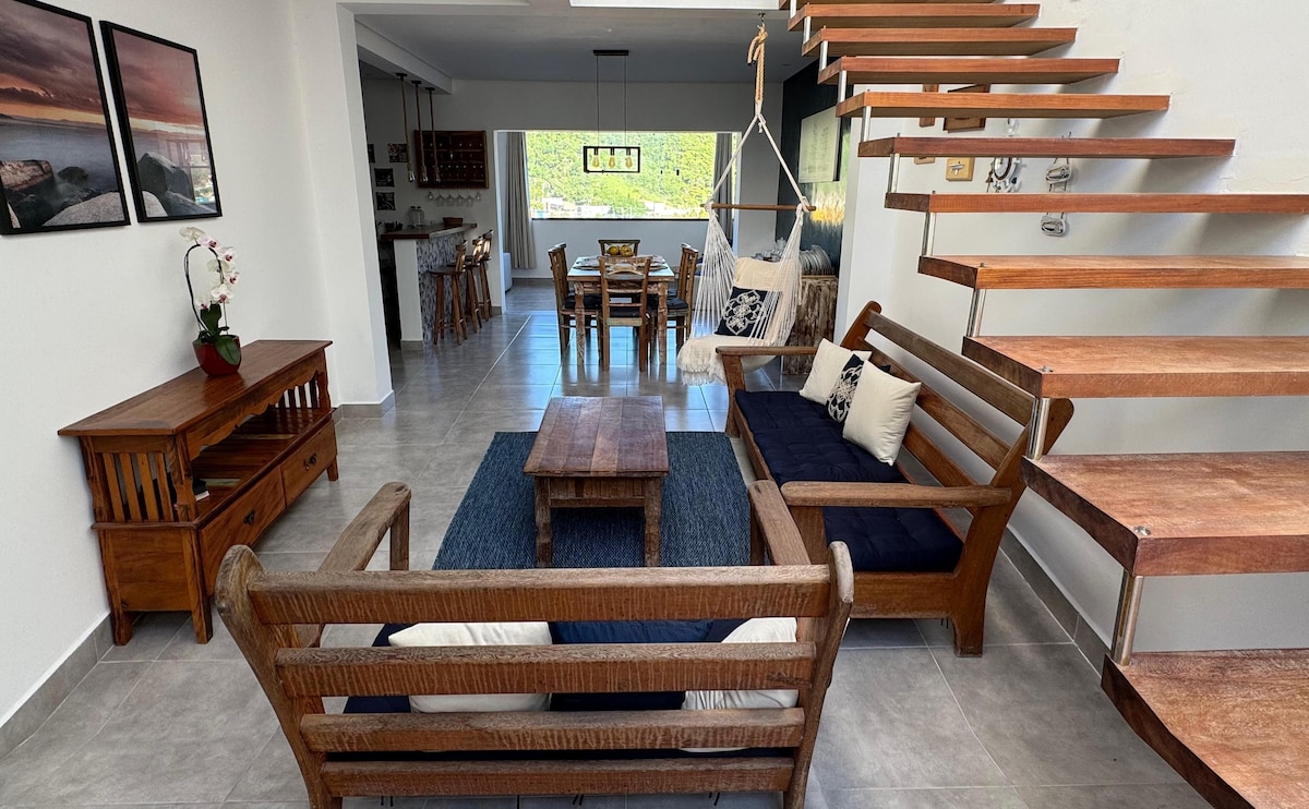 An open living space features wooden furniture, including a coffee table and two seating areas with navy cushions. A staircase made of wood leads to the upper level, while a dining area with wooden chairs is visible in the background.