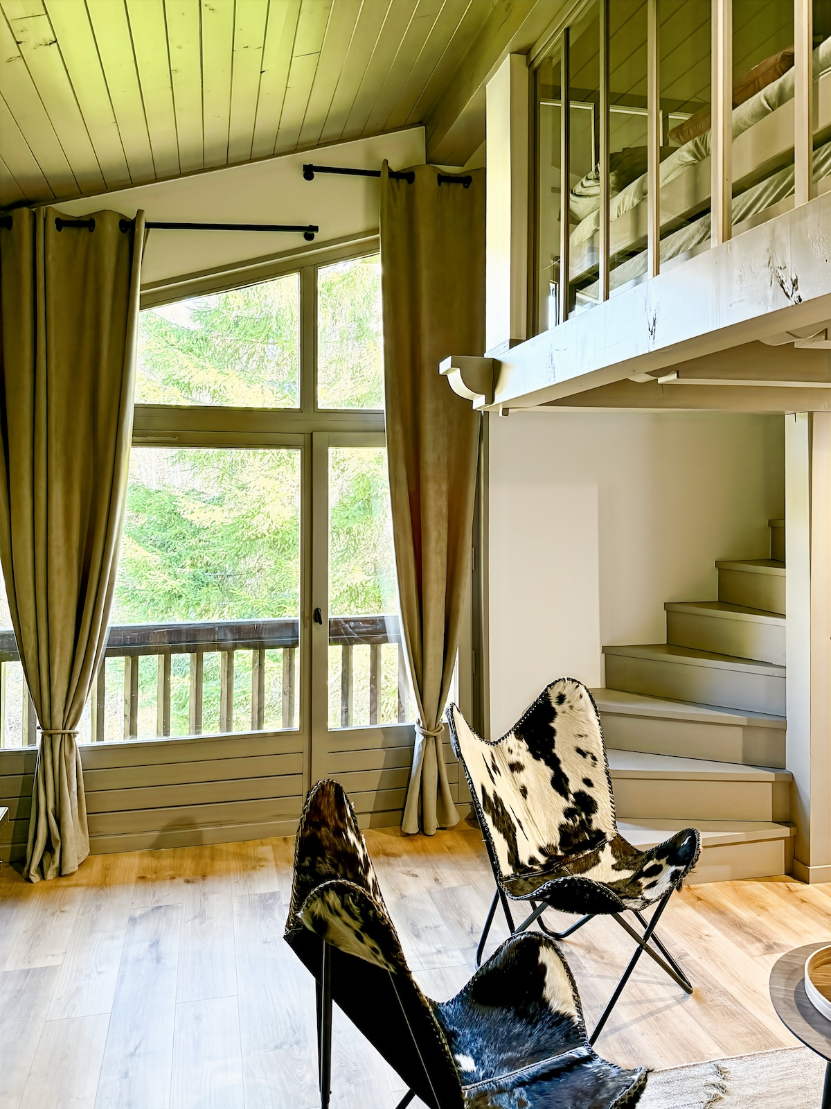 A light-filled living space features large windows offering views of the surrounding trees. Two unique chairs with cowhide accents are positioned on the wooden floor. A staircase leads to a mezzanine area, enhancing the open layout of the apartment.