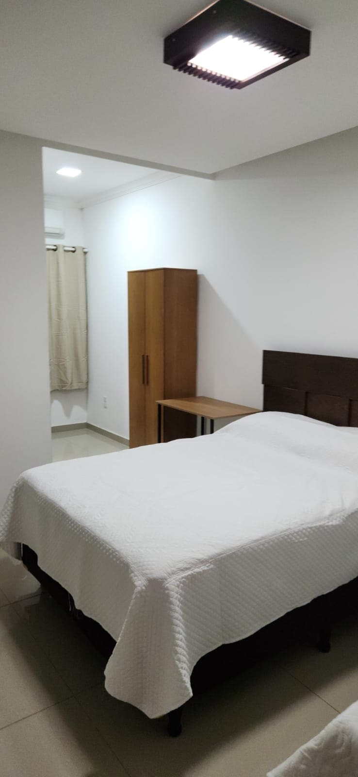 A simple and modern sleeping area features a spacious bed with a light-colored coverlet. A wooden wardrobe and a small desk are positioned against the walls. A window is partially covered by a light curtain, allowing natural light to gently illuminate the space.