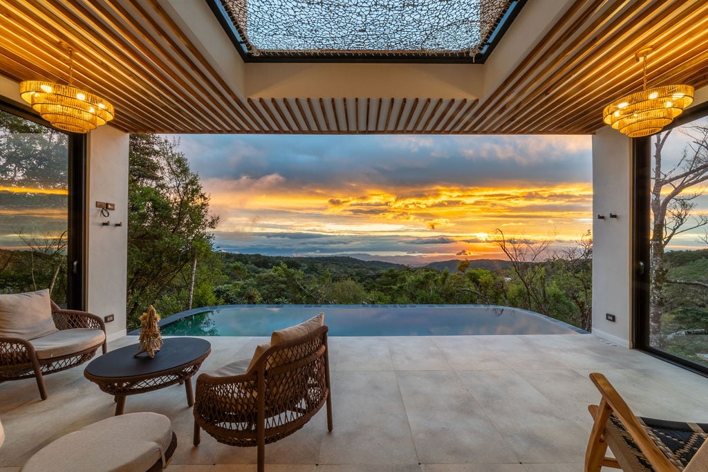A serene outdoor living space features comfortable seating arranged around a low table, with panoramic views of a sunset over lush greenery. An infinity pool reflects the vibrant colors of the sky, framed by large glass doors that open to the natural surroundings.