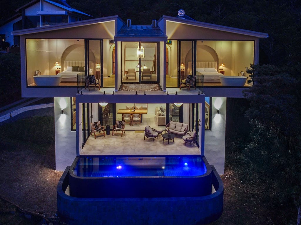 The exterior view captures a modern architectural design with two levels featuring large glass panels. The structure hovers over a serene infinity pool illuminated in blue, surrounded by a natural setting. Soft lighting highlights the elegant furnishings on the terrace below.