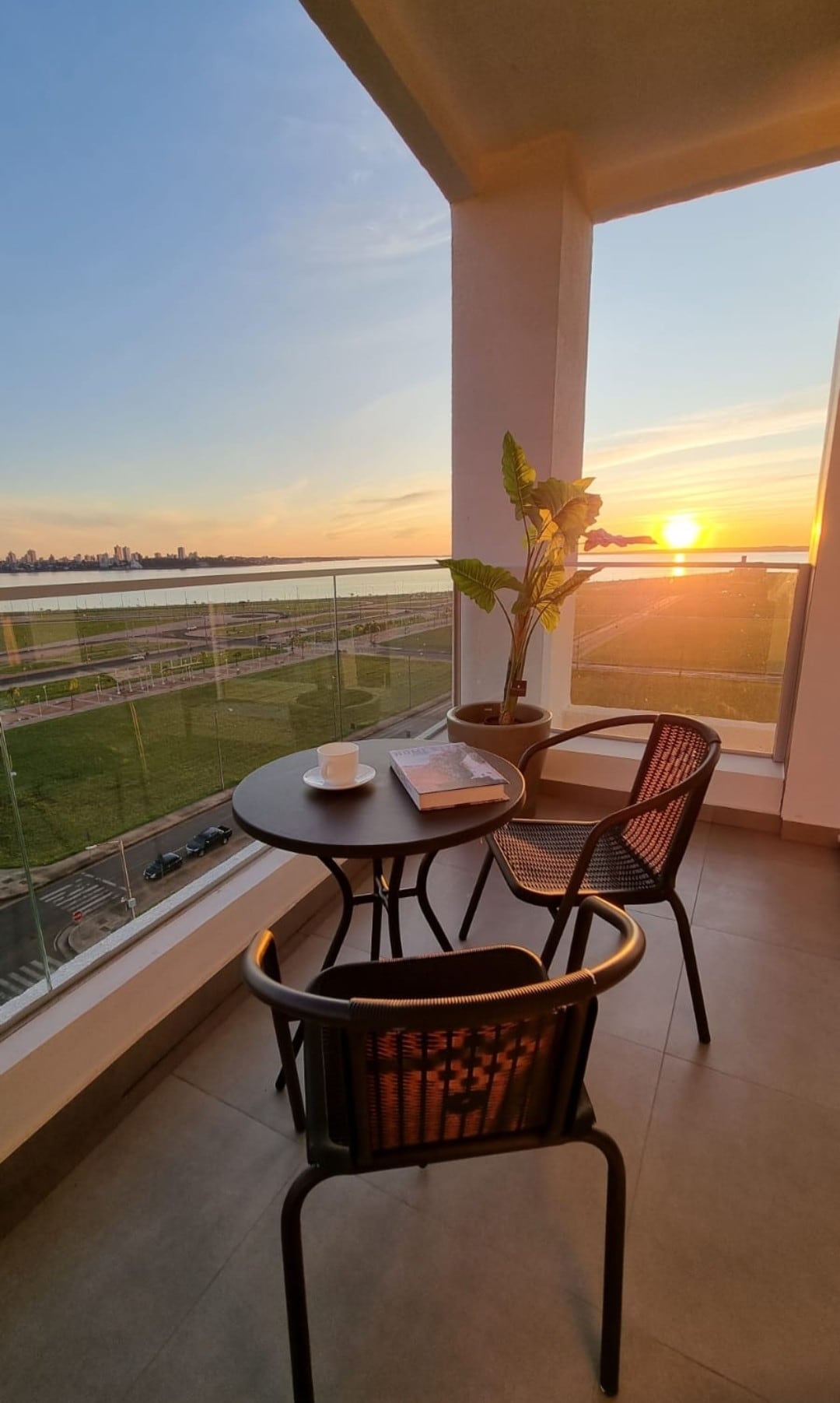 A balcony setting features a small round table accompanied by two chairs. A cup rests on the table, while a potted plant adds a touch of greenery. The scene is illuminated by the warm glow of a sunset, with distant city views visible beyond.