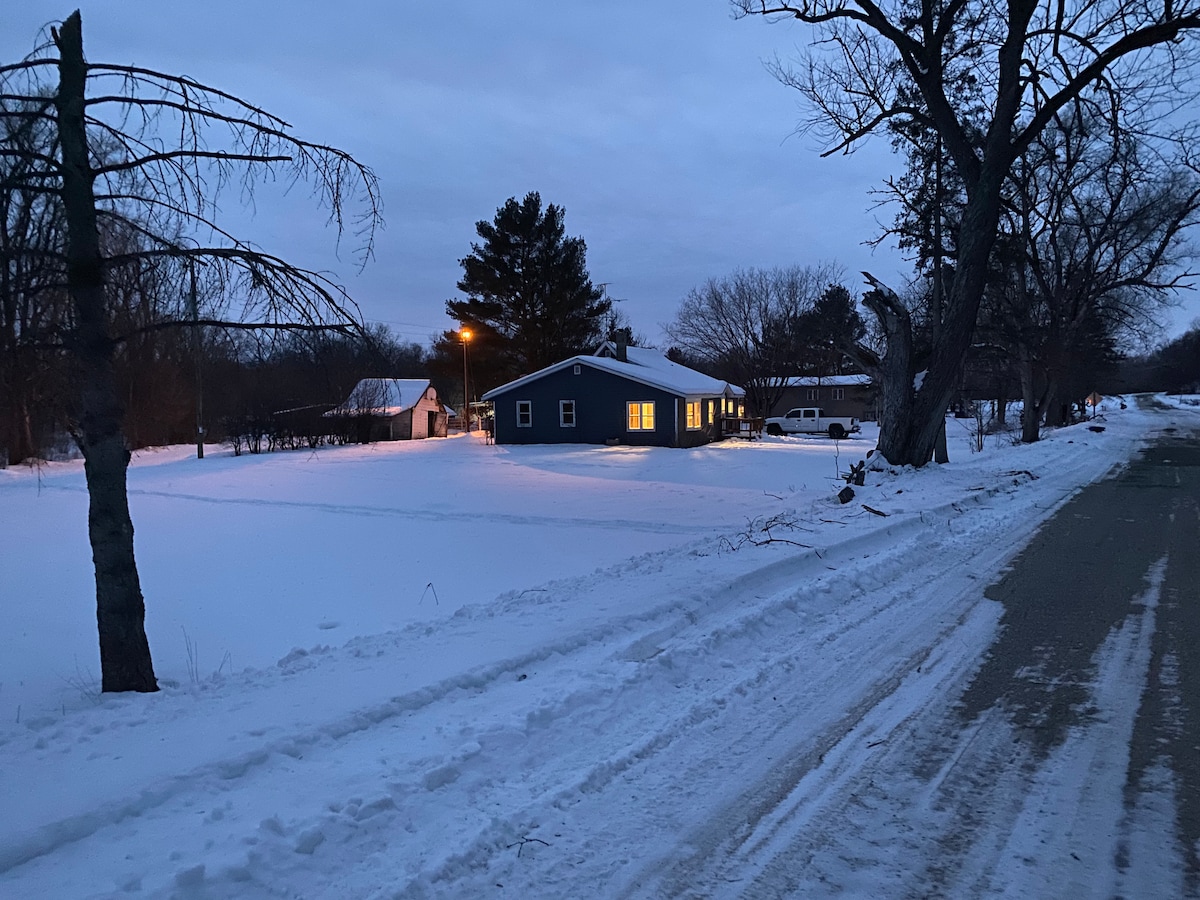 A blue home is visible from the street, illuminated warmly from within. Snow blankets the ground, reflecting the soft light of a streetlamp. Surrounding trees and open space create a serene winter landscape, emphasizing the tranquil setting of the property.
