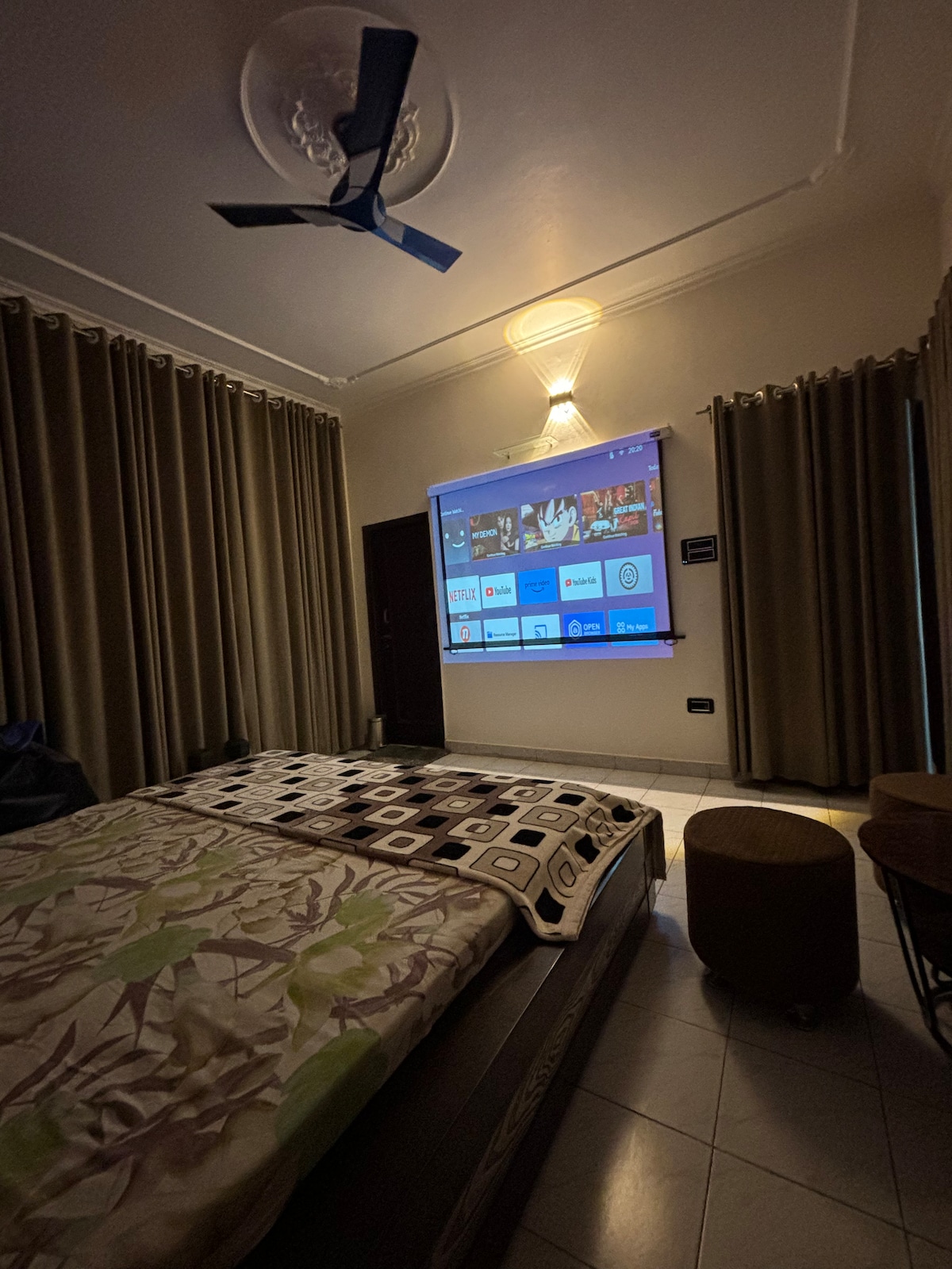 A private room features a spacious bed covered with a patterned blanket, while a projector screen is mounted on the wall, displaying various streaming options. Soft lighting is provided by a ceiling fixture, and long curtains frame a window, enhancing the room's cozy ambiance.