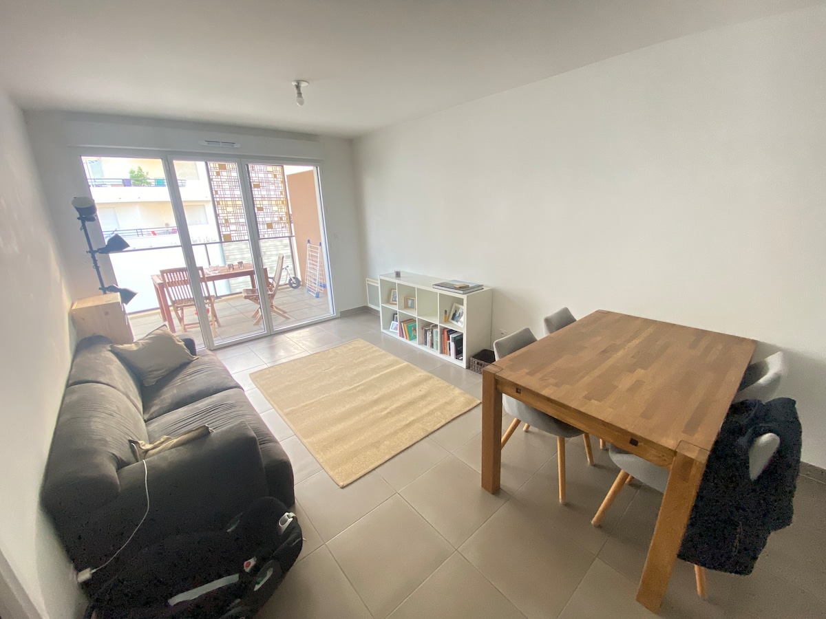 An airy living space is presented, featuring a dark sofa and a wooden dining table surrounded by light-colored chairs. A carpet is positioned on the tiled floor, while a large sliding door opens to a balcony, allowing natural light to fill the room.
