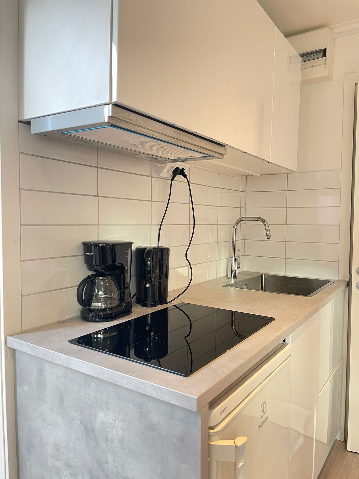 The kitchen area features modern cabinetry and a sleek countertop. A coffee maker and kettle are positioned beside a sink, while an electric stovetop is built into the surface. A refrigerator is discreetly placed underneath the counter, enhancing functionality.