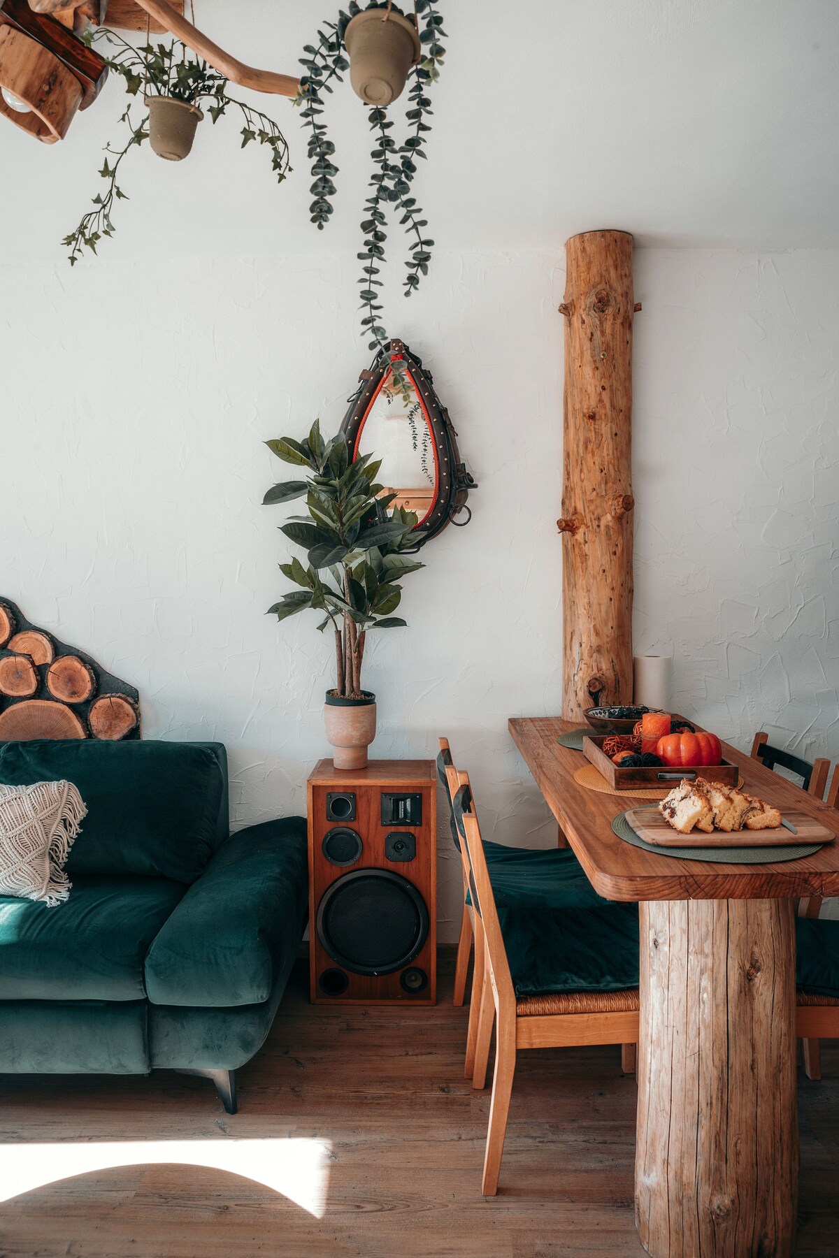 A welcoming living space features a green sofa placed next to a wooden table surrounded by chairs. A potted plant stands near an ornate mirror, while natural wood accents add to the rustic feel. A wooden log wall complements the character of the room.