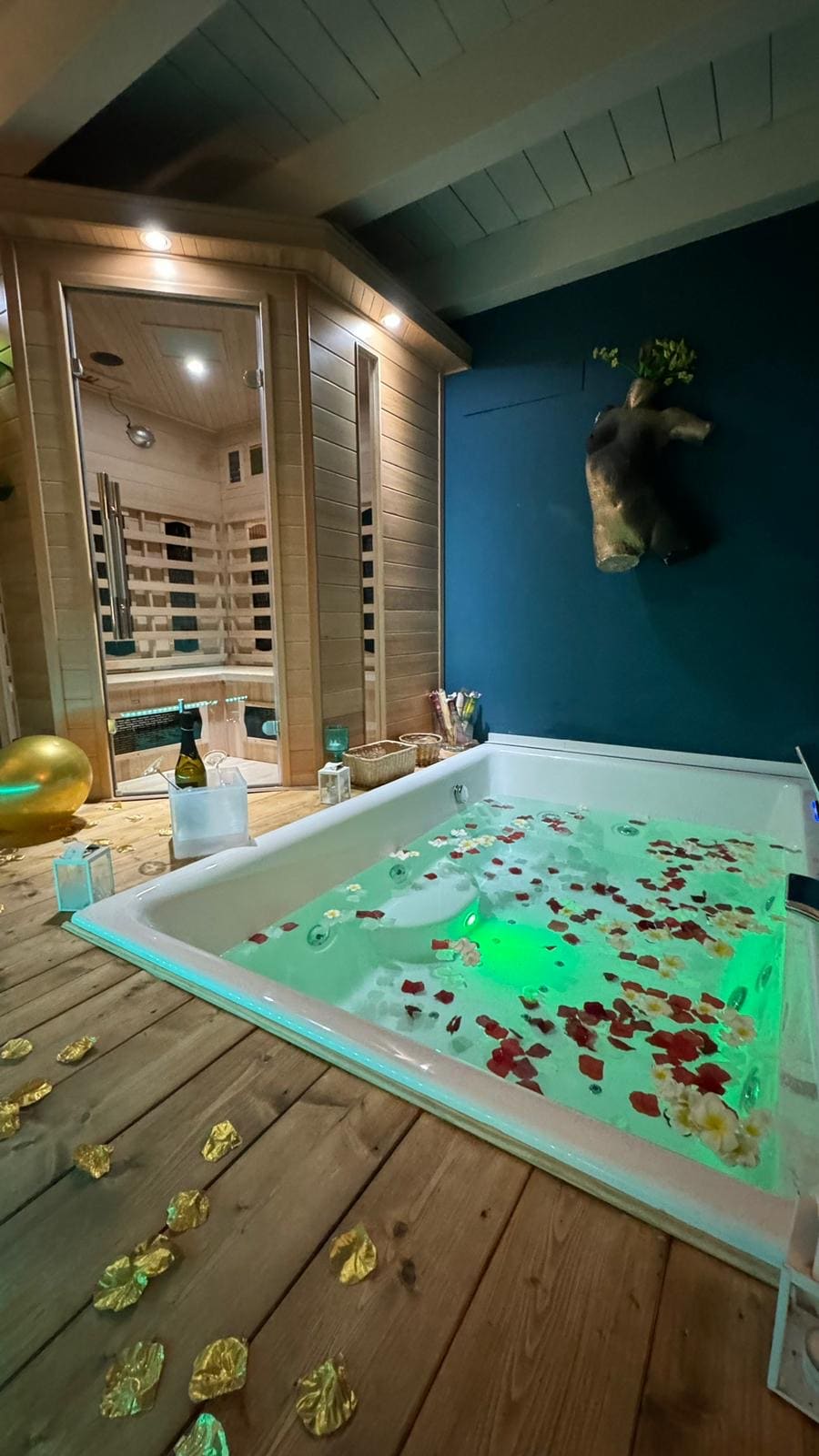 A private spa area features a large Jacuzzi adorned with rose petals, set against a backdrop of a wooden sauna. Soft green lighting highlights the space, while decorative elements create a serene atmosphere. The wooden floor adds warmth to the intimate setting.