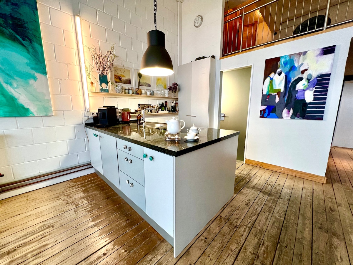 A spacious kitchen area features modern cabinetry and a sleek black countertop with various appliances. Above, a large pendant light illuminates the space. Artworks adorn the walls, and wooden flooring adds warmth to the environment, leading towards an adjacent open space.