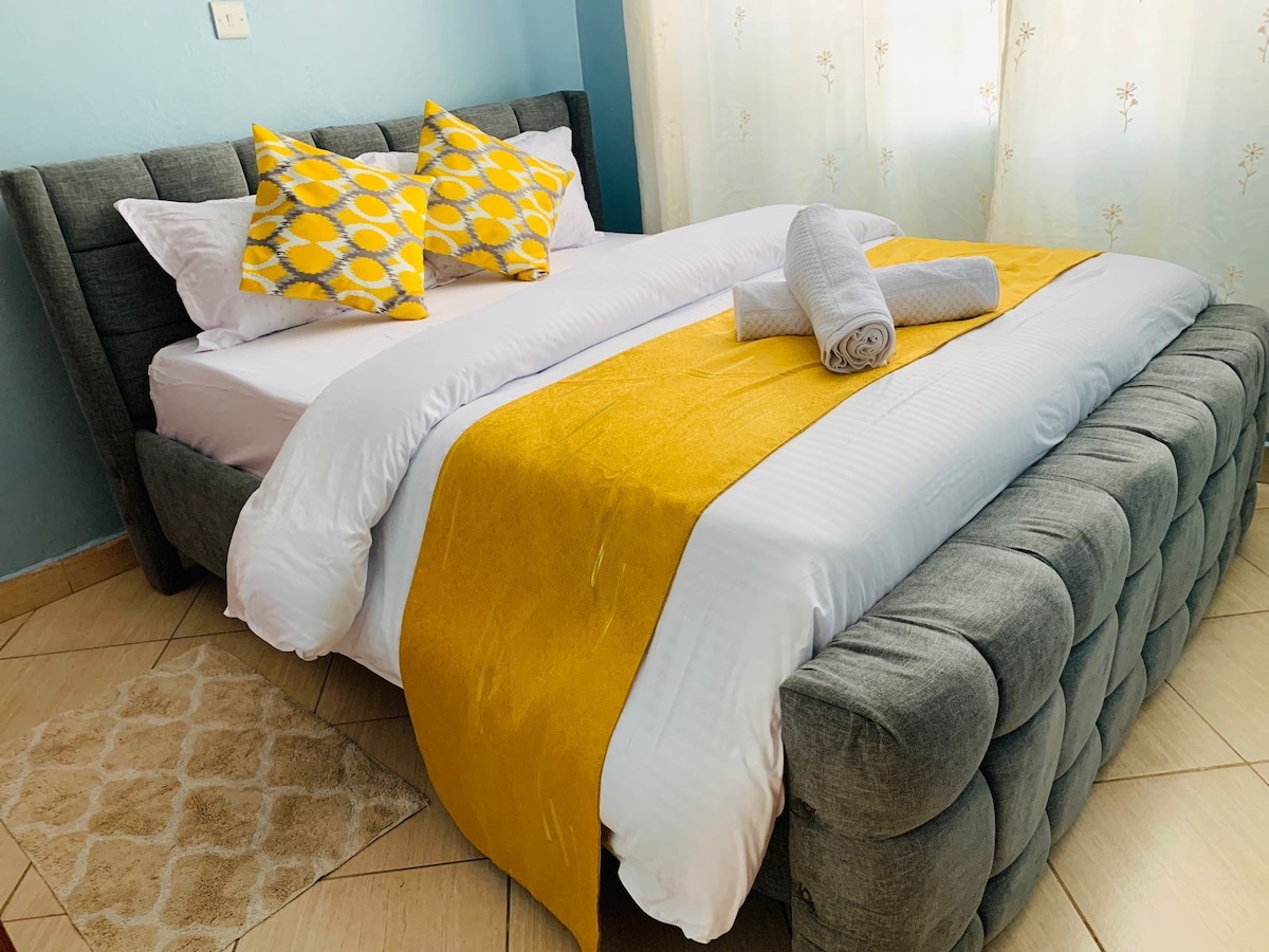 A spacious bed is centered in the room, adorned with a bright yellow throw and decorative pillows featuring a geometric pattern. Fresh white linens complement the grey upholstered headboard. Soft towels are neatly rolled on the bed, and light filters through sheer curtains.