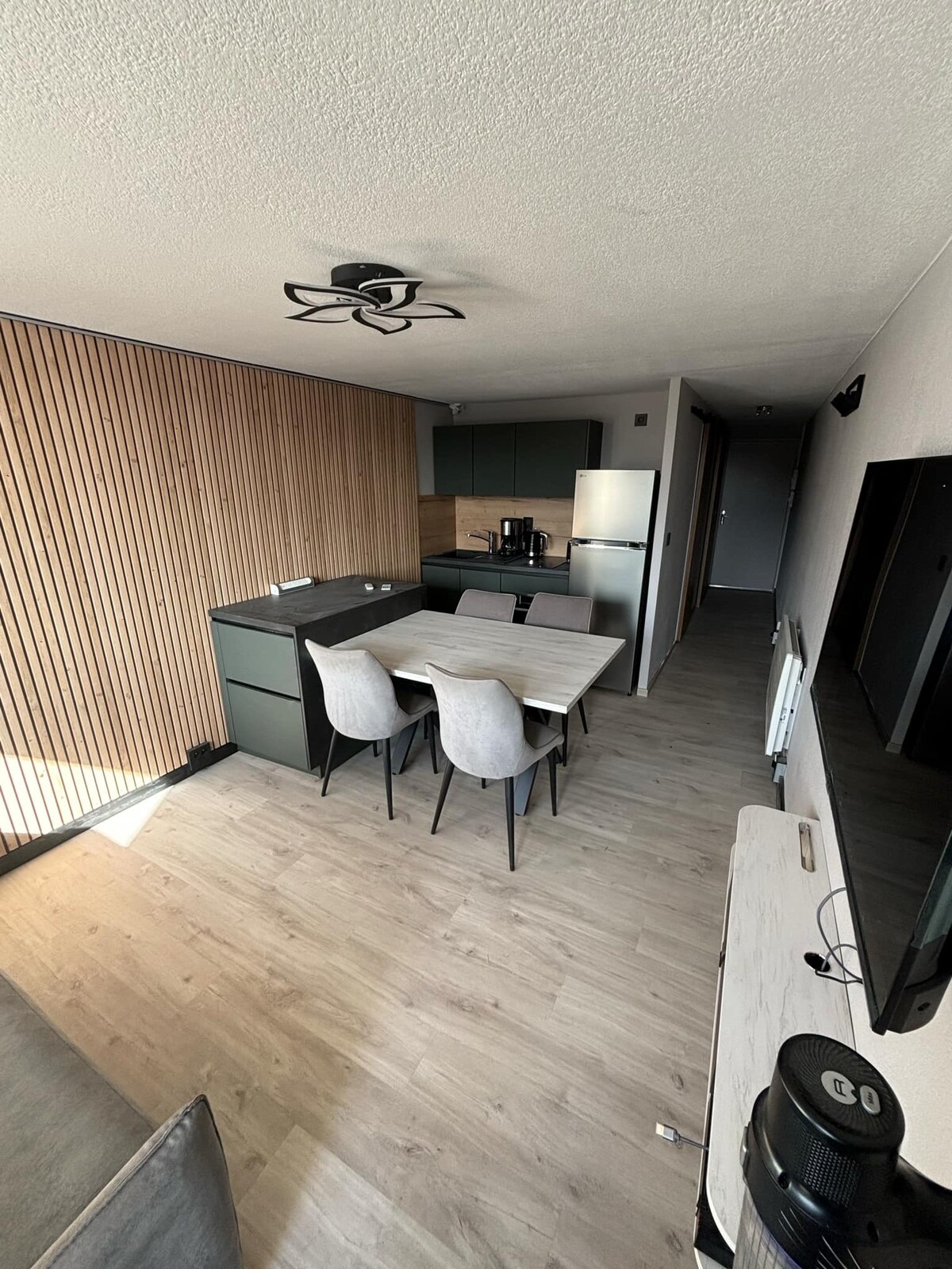 A modern kitchen and dining area are visible, featuring a sleek wooden dining table surrounded by four chairs. The kitchen includes minimalist cabinetry and appliances, with natural light illuminating the space through a nearby window. Light wood flooring complements the warm wooden paneling on the wall.
