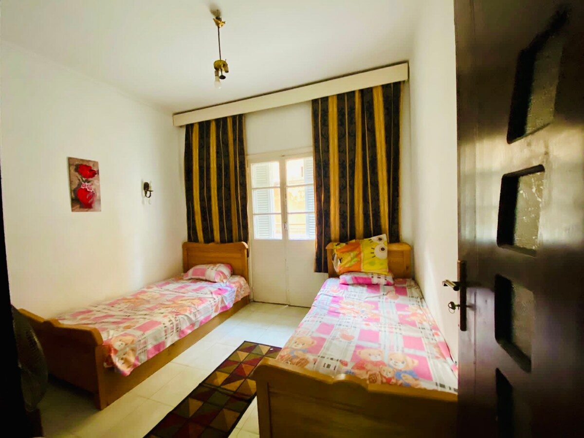 A simple bedroom features two beds with colorful bedding set against light-colored walls. Natural light streams through a door leading to an outdoor area, which is adorned with striped curtains. A piece of wall art with floral motifs adds a touch of decor.