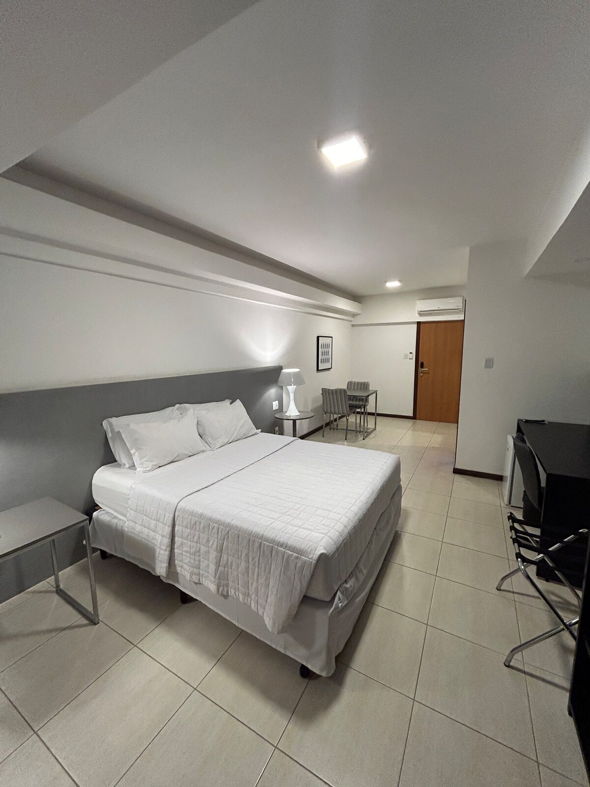 A well-lit loft features a spacious bed dressed in white linens, flanked by two bedside lamps. A work desk is positioned next to a chair, and the floor is tiled in neutral tones, enhancing the airy atmosphere. The entrance door is visible in the background.