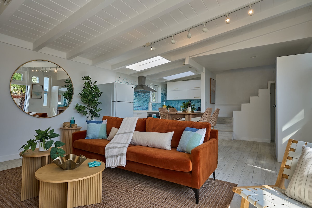 A spacious living area features a vibrant orange sofa adorned with soft pillows. A round mirror hangs above a wooden coffee table with decorative items. Natural light pours in through large windows, illuminating the inviting open concept design leading to a dining area and kitchen.