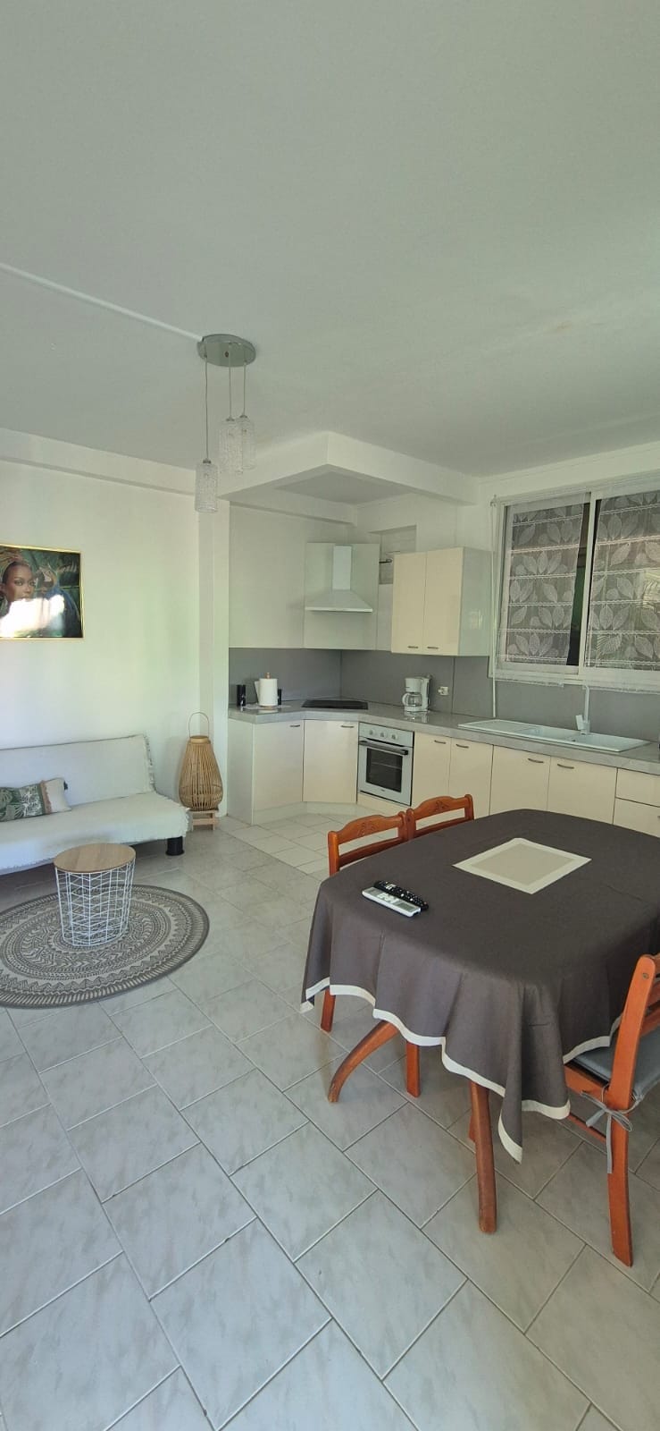 A well-lit kitchen and dining area feature light cabinetry and modern appliances. A round table is set with a dark tablecloth and four wooden chairs. A comfortable seating area is seen nearby, complemented by a circular rug and a decorative wall hanging.