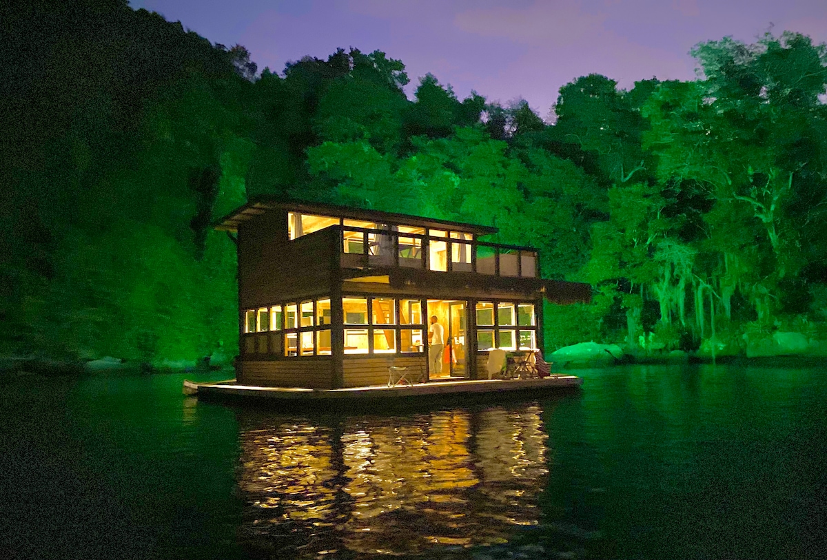 The houseboat is illuminated at night, revealing its modern design and large windows. Surrounded by lush greenery, its reflective surface in the calm water enhances the serene atmosphere. The structure's exterior showcases an inviting blend of wood and glass against the darkened backdrop of nature.