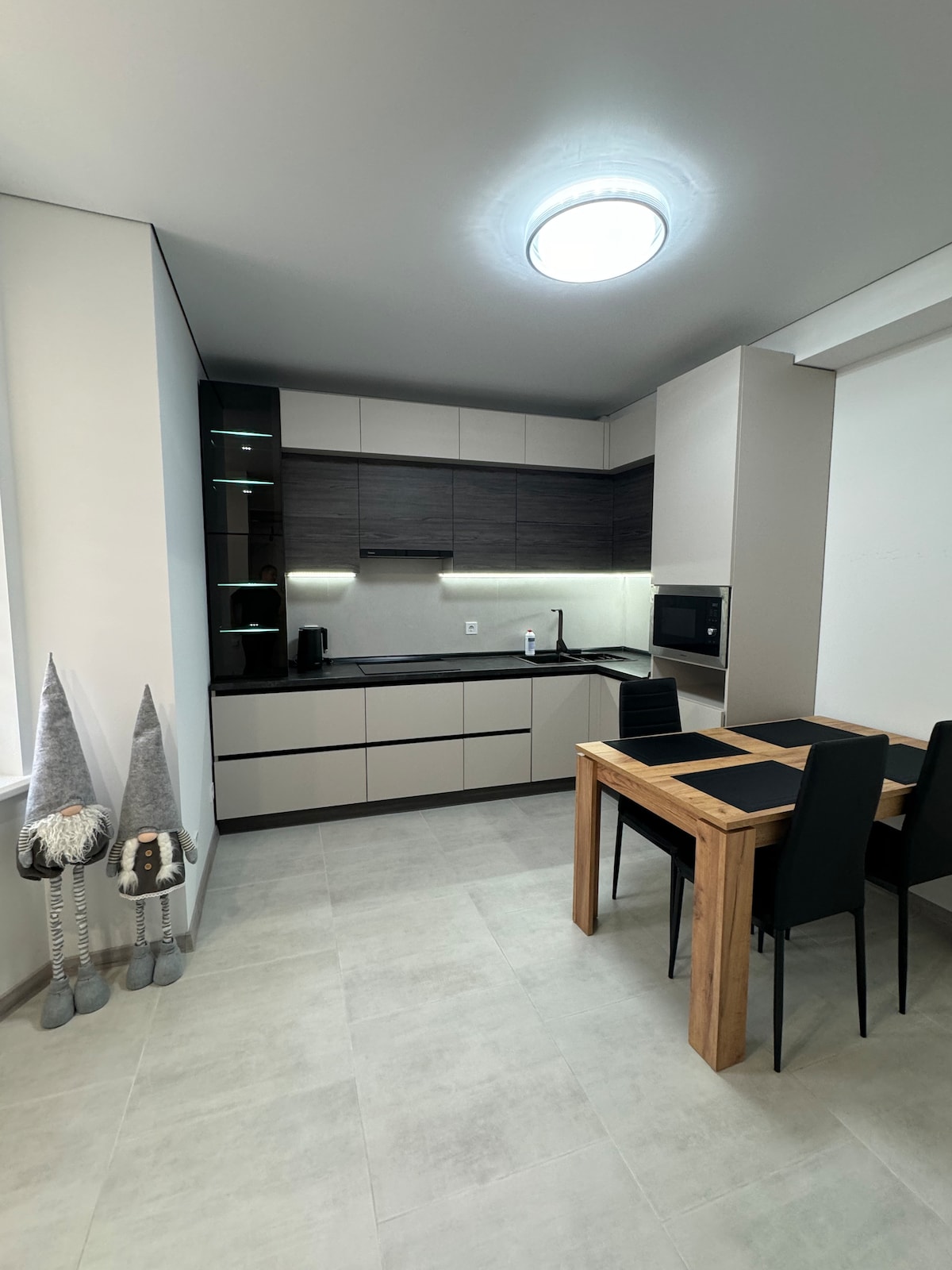 A modern kitchen is designed with sleek cabinetry and contrasting dark wood accents. Counter space is provided alongside built-in appliances. A small wooden dining table is surrounded by four black chairs, and two decorative figures stand next to the window.