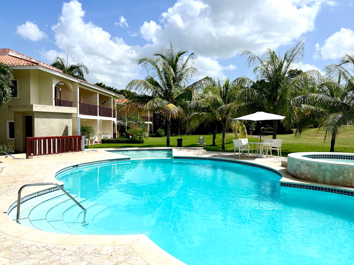 A serene pool area features a large, curved swimming pool surrounded by palm trees. A jacuzzi is positioned adjacent to the pool, with outdoor seating provided under a white umbrella. The two-story building is visible in the background, complemented by greenery and a clear blue sky.
