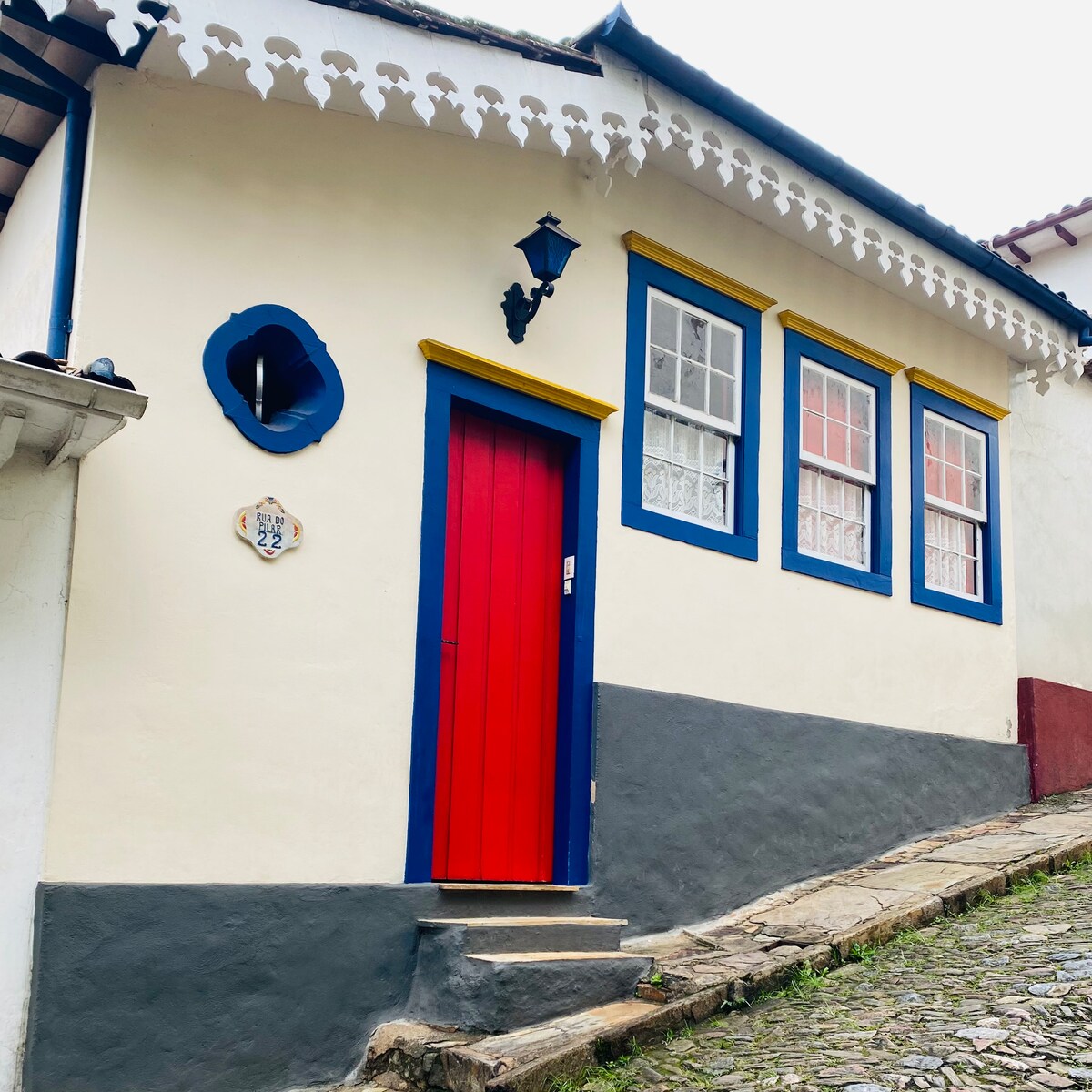 The exterior of the charming house features a bright red door framed by blue trim. Three windows with lace curtains invite natural light, while decorative details enhance the entrance. The building's white walls and the unique sloped street create a picturesque setting.