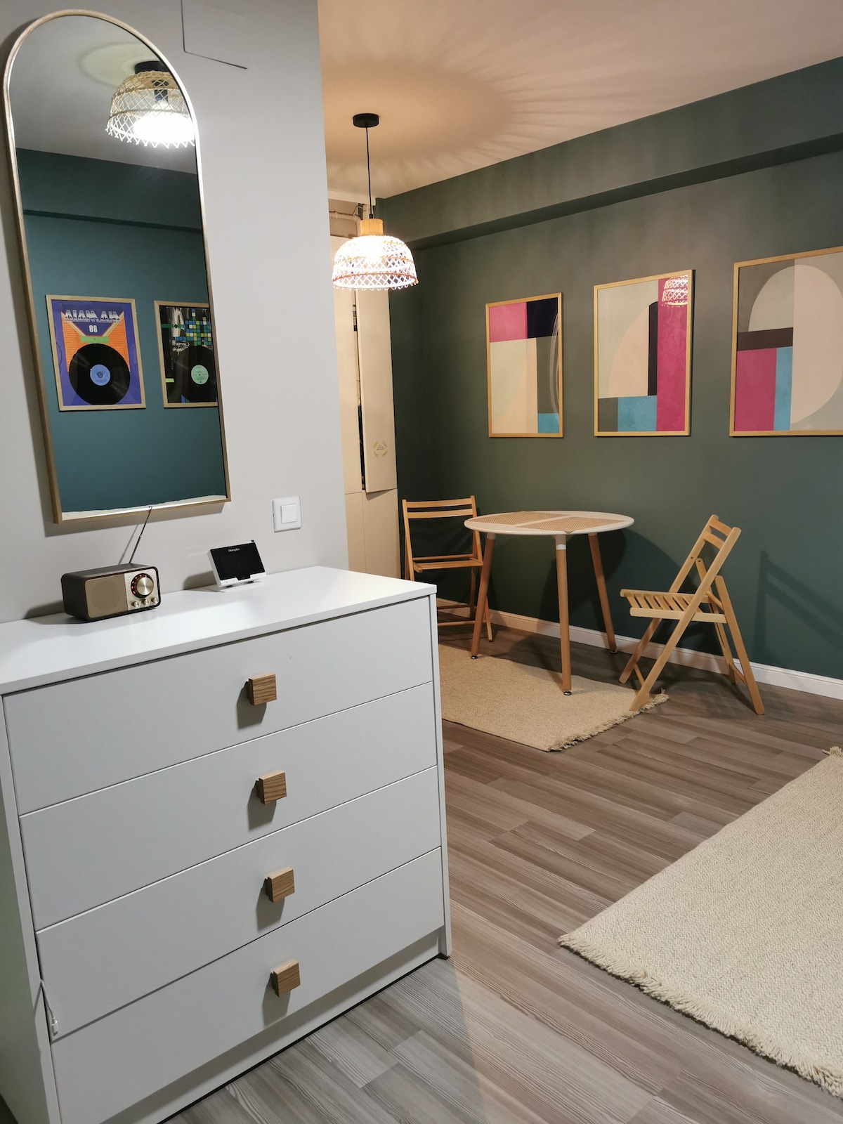 A well-organized living space is highlighted by a stylish mirror above a white dresser. Artwork in various colors adorns the green walls. A small round table is accompanied by two wooden chairs, creating a cozy dining area. Warm lighting adds a relaxed ambiance.