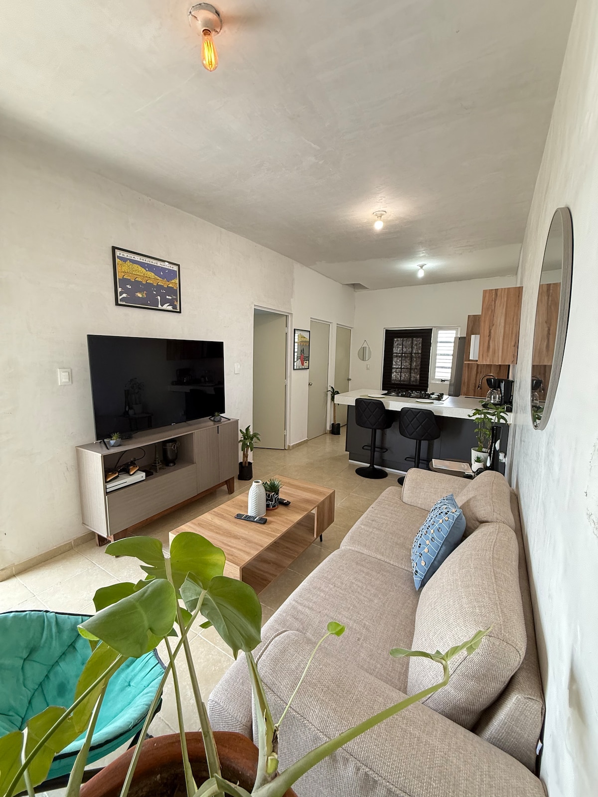 A living area features a light-colored sofa set against a textured wall, complemented by a coffee table. A television is mounted on the wall, and two bar stools sit easily at a counter. Natural light enters through an adjacent doorway, enhancing the serene environment.