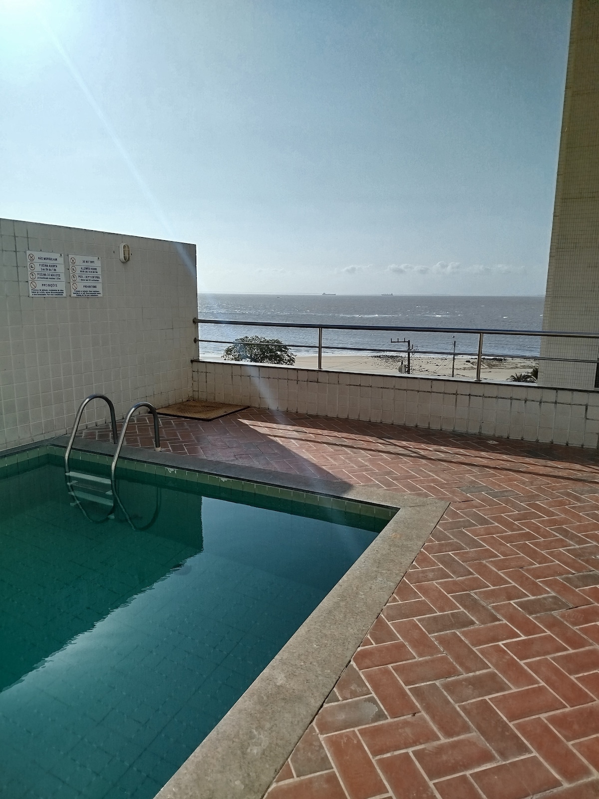 A clear view of the swimming pool reflects the sky above, with steps leading into the water. Beyond the pool, the beach is visible, framed by a railing and distant shoreline. Sunlight casts warm tones across the tiled deck.