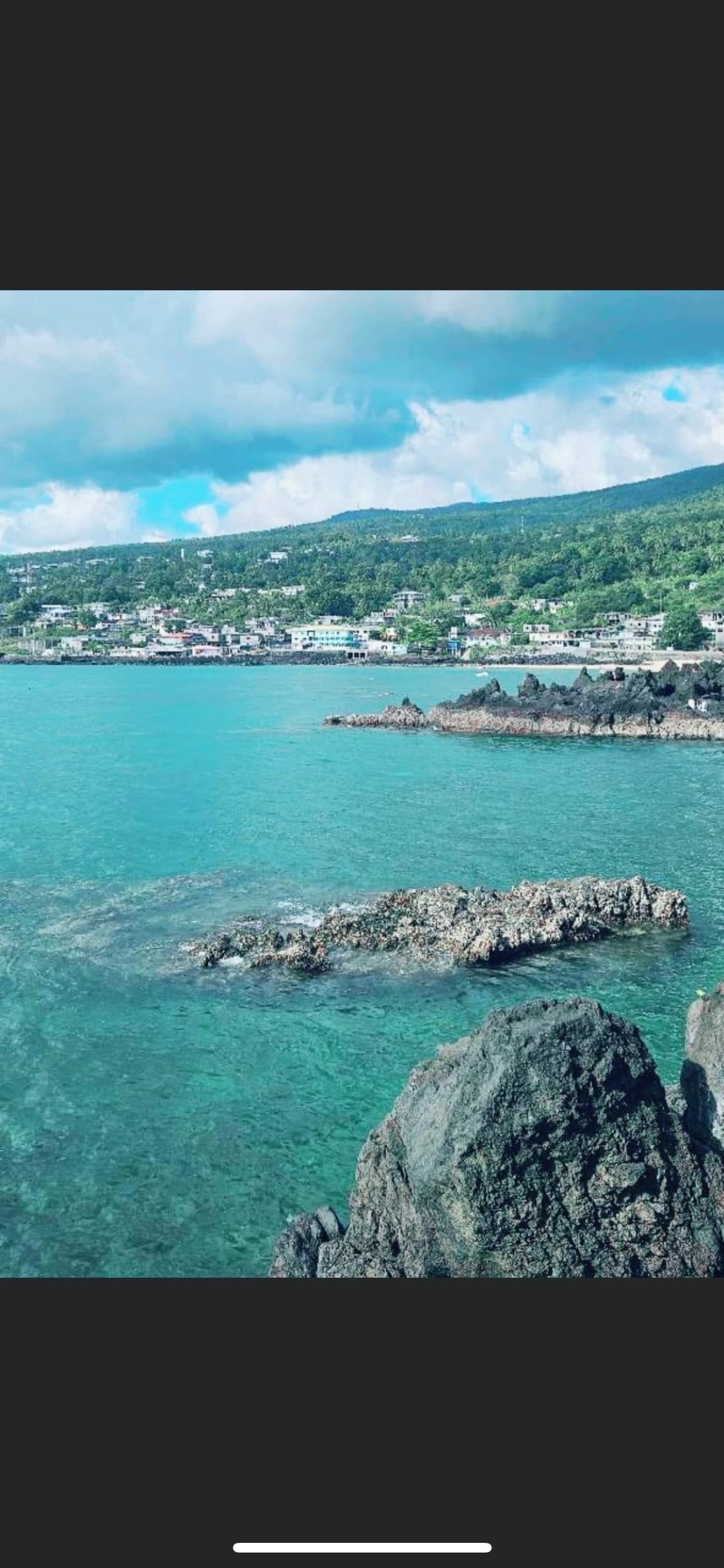 A coastal view showcases clear blue waters gently meeting rocky shores. Lush green hills rise in the background, lined with quaint buildings, enhancing the serene ambiance of the landscape.
