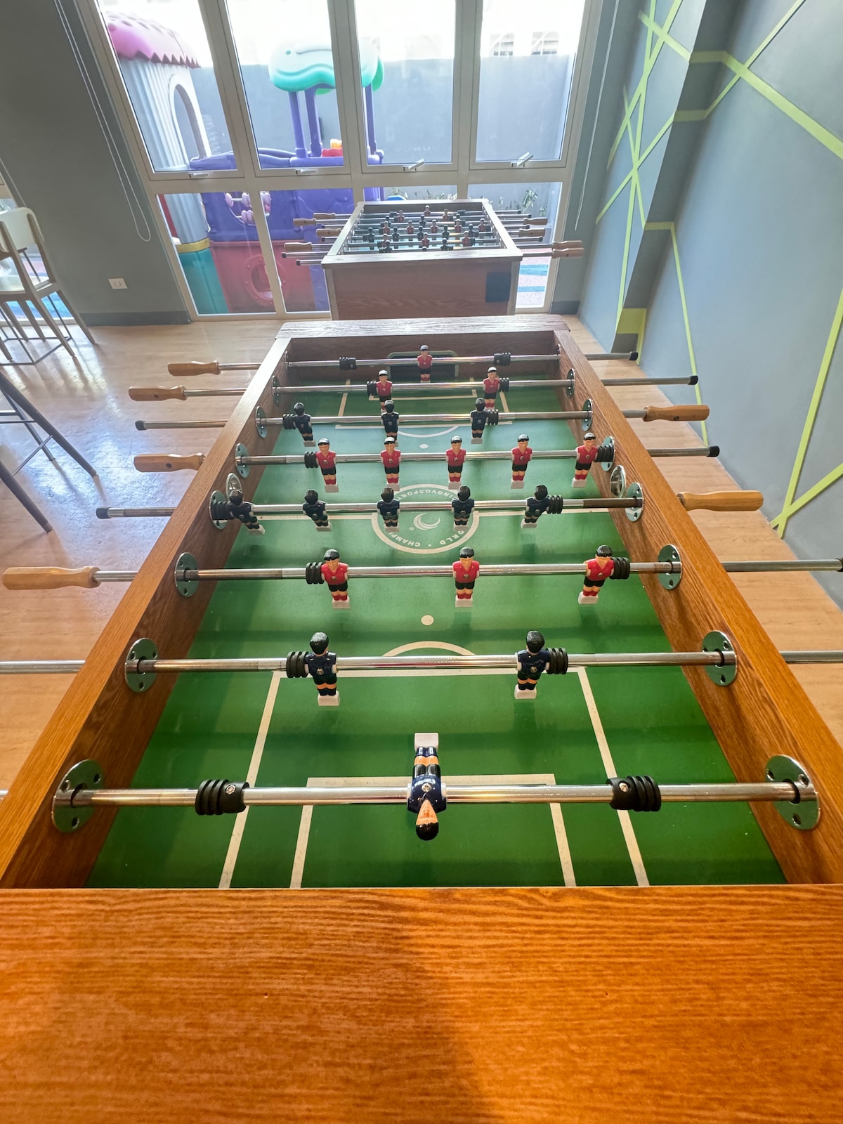 A foosball table is featured, with players arranged in two teams on a green playing field. The wooden frame and handles add a classic touch, while large windows in the background allow natural light to illuminate the space.