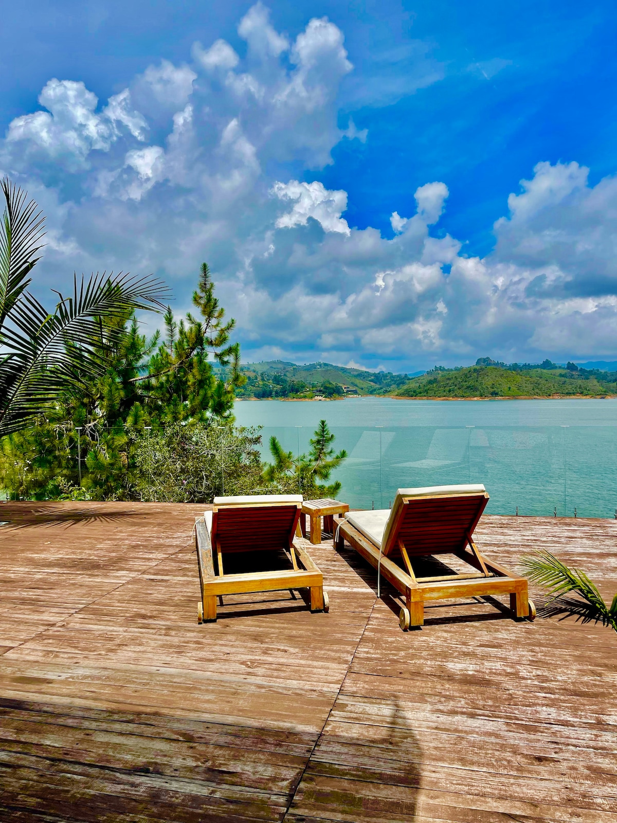 Two wooden lounge chairs are positioned on a spacious deck overlooking a serene body of water. Lush greenery and distant hills create a natural backdrop, with a bright blue sky dotted with clouds enriching the tranquil setting.